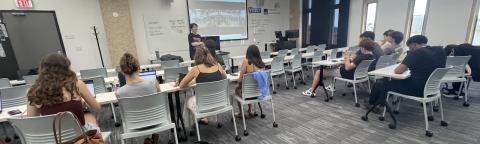 Students seated in a classroom, with a presenter standing by a projection screen.
